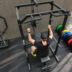 A man in a black t-shirt and shorts sits on a bench inside a black power rack, pulling a bar with a barbell. The gym floor is black rubber matting, and weight plates in blue, green, yellow, and red are stacked on the right against a slatted wood wall.
