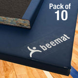 Close-up of a blue gym mat with 'beemat' branding, showing a cut corner revealing multicolored foam. Text in the corner reads 'Pack of 10'. The mat rests on a wooden floor.