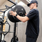 A man in a black t-shirt and black cap adjusts a barbell with black weight plates on a power rack in a gym setting.