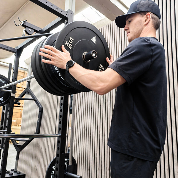 A man in a black t-shirt and black cap adjusts a barbell with black weight plates on a power rack in a gym setting.
