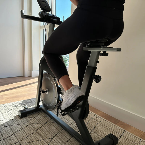A person riding a HIT Fitness spin bike, with a city view through the window. The rider wears black workout gear and white sneakers.