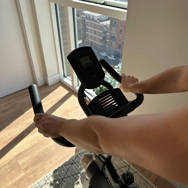 The image shows a close-up of a person’s hands gripping the handlebars of a HIT Fitness spin bike, with the bike's console visible. The display shows workout data, including speed, while a city view is seen through the window.