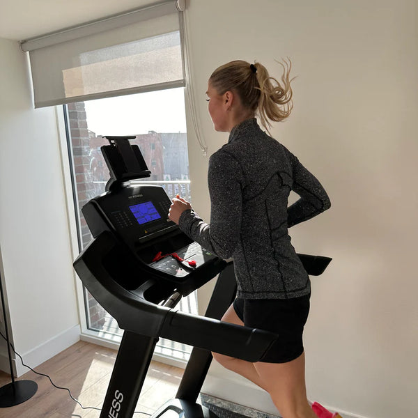 A woman running on a HIT Fitness treadmill, wearing a gray jacket and pink sneakers, with a tablet mounted on the console.