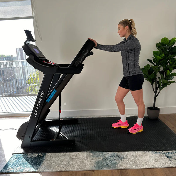 A woman using a HIT Fitness treadmill, standing beside it while holding the console, wearing athletic gear and pink running shoes.
