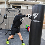 A man in a black hoodie and shorts practices boxing in an indoor gym, punching a large black HIT BOXING heavy bag. Weight plates are stacked on a rack in the background.