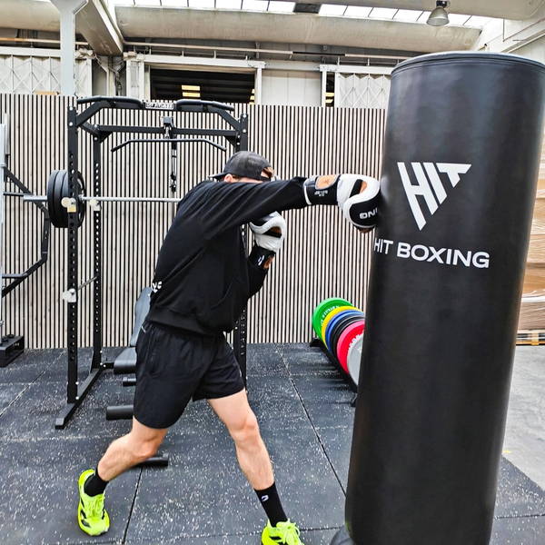 A man in a black hoodie and shorts practices boxing in an indoor gym, punching a large black HIT BOXING heavy bag. Weight plates are stacked on a rack in the background.