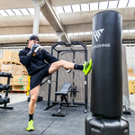 A man in a gym kicks a black heavy punching bag. The punching bag has a white logo that reads 'HT BOXING'.