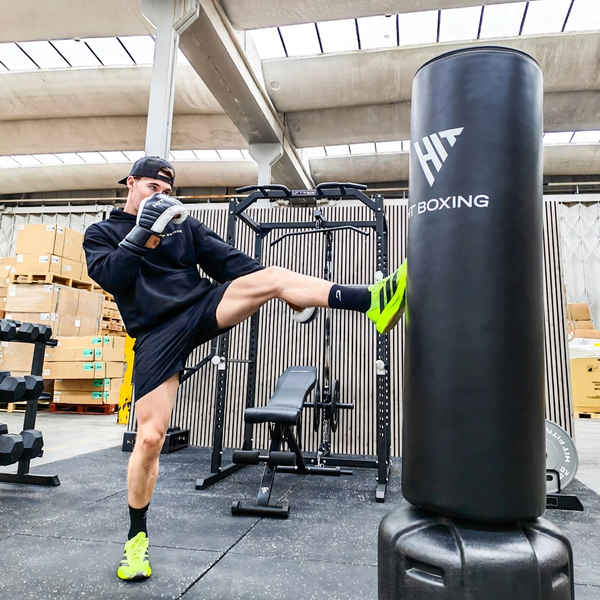 A man in a gym kicks a black heavy punching bag. The punching bag has a white logo that reads 'HT BOXING'.