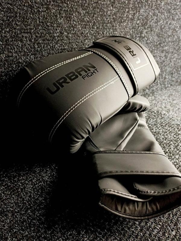 A pair of black Urban Fight punch bag mitts with white stitching and velcro straps, placed on a dark textured surface with soft lighting.