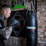 A tattooed man wearing black Urban Fight punch bag mitts is in a boxing stance facing a hanging black Urban Fight punching bag in a gym with brick walls and wall art in the background.