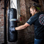 A man wearing black Urban Fight punch bag mitts punches a hanging black Urban Fight punching bag in a gym with brick walls, a window, and a red trophy on a shelf in the background.