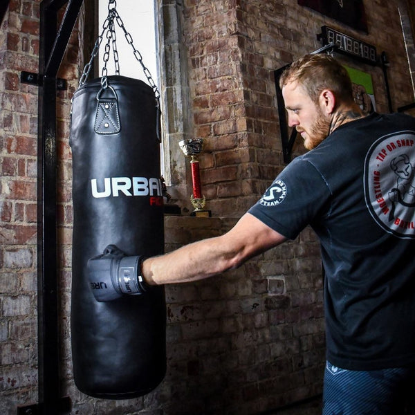 A man wearing black Urban Fight punch bag mitts punches a hanging black Urban Fight punching bag in a gym with brick walls, a window, and a red trophy on a shelf in the background.