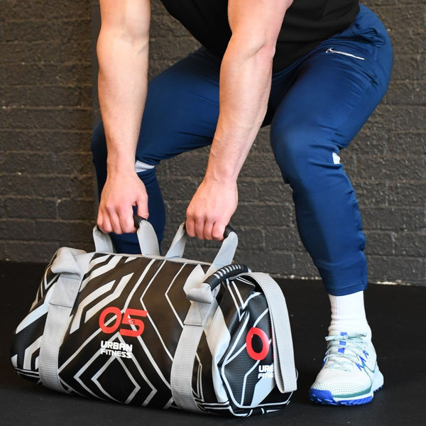 Person performing a deadlift with an Urban Fitness Power Bag 5kg, featuring ergonomic handles and straps, set against a plain gym wall background.