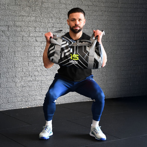 Person performing a squat with an Urban Fitness Power Bag 15kg, featuring ergonomic handles and straps, set against a plain gym gray wall background.