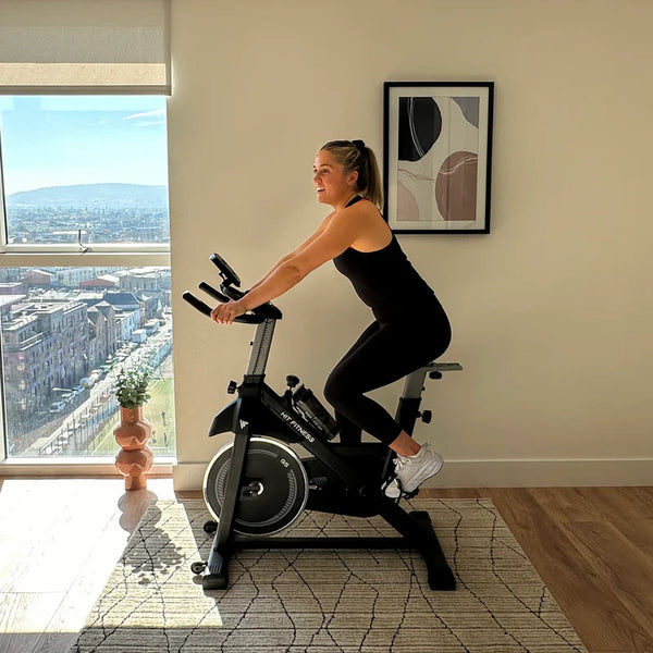 The image shows a woman using a HIT Fitness spin bike in a bright, modern room. She is dressed in athletic wear and riding the bike with a view of the city through the window. The bike features a large flywheel and adjustable handlebars.